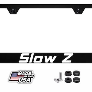 May include: A black rectangular number plate frame featuring the white text Slow Z printed on the bottom edge. The set includes two metal screws, four black rubber washers, and four black screw covers, along with a Made in the USA emblem.