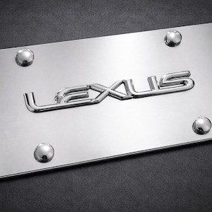 Puede incluir: Un marco de matrícula rectangular de cromo pulido con la palabra LEXUS en relieve metálico en el centro. El marco incluye cuatro cubiertas de tornillos de montaje redondeadas de tono plateado situadas en cada esquina.