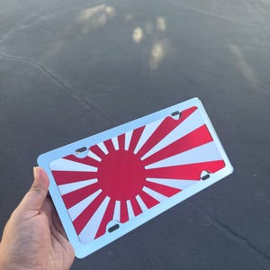 Puede incluir: Placa de matrícula de coche con un diseño de sol rojo y rayos blancos sobre fondo blanco. La placa tiene un borde azul claro. El diseño recuerda a la bandera imperial japonesa, sostenida en una mano.