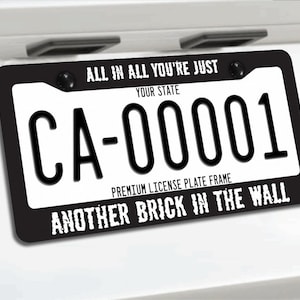 Op de afbeelding: Zwarte kentekenplaathouder met witte letters. De tekst "ALL IN ALL YOU'RE JUST" staat bovenaan, met "CA-00001" en "ANOTHER BRICK IN THE WALL" onderaan. De houder is gemonteerd op een wit oppervlak.