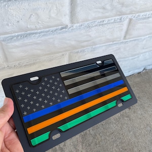 Może przedstawiać: Czarna ramka na tablicę rejestracyjną z wystylizowanym wzorem amerykańskiej flagi. Flaga zawiera niebieskie, pomarańczowe i zielone paski z polem gwiazd. Ramka jest trzymana na tle białej ceglanej ściany.