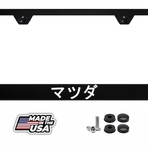 May include: A black rectangular license plate frame featuring the Japanese text "マツダ" in white on the bottom edge. The set includes two metal screws and four black plastic screw covers, accompanied by a "MADE IN THE USA" graphic with a flag.