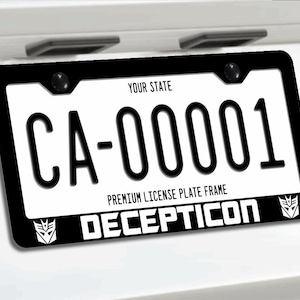 Op de afbeelding: Een zwarte kentekenplaathouder met het woord DECEPTICON in witte hoofdletters aan de onderkant, geflankeerd door twee witte Decepticon-logo's. Het frame omringt een witte kentekenplaat met de tekst YOUR STATE en de code CA-00001.
