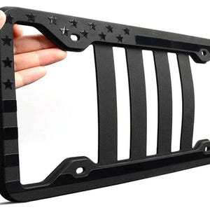 May include: Black license plate frame with a design featuring stars and stripes. The frame has a matte finish and a rectangular shape with vertical bars and star accents. The frame is held by a hand.