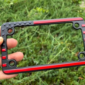 May include: Black license plate frame with a red stripe and accents. The top left corner features a design of stars. The frame has mounting holes and is held in a hand against a green background.