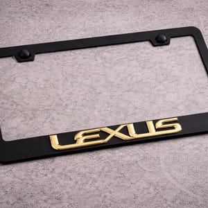 Puede incluir: Un marco de matrícula rectangular negro que presenta la palabra LEXUS en letras doradas en relieve centradas a lo largo del borde inferior. El marco incluye dos tapas de montaje negras circulares en las esquinas superiores.