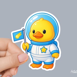 May include: A sticker featuring a cartoon duck in a white and blue astronaut suit, holding a flag with a yellow comet design. The duck has a yellow body, orange beak, and is wearing a blue helmet. The sticker is held by a hand.