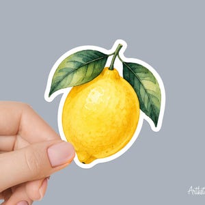 May include: A die-cut sticker featuring a vibrant yellow lemon with a textured, watercolor-style finish. The lemon is attached to a short green stem with two detailed, dark green leaves extending outward, all outlined in a clean white border.