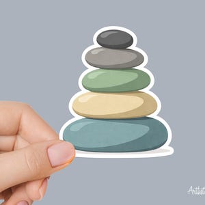 May include: A sticker featuring a stack of six smooth, colorful stones in shades of blue, yellow, green, and gray. The stones are stacked on top of each other, creating a balanced tower. The sticker has a white border and is held by a hand.