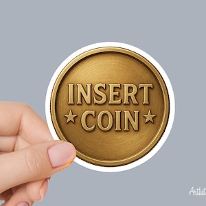 May include: A round sticker with a gold coin design. The coin features the words "INSERT COIN" in bold, capital letters, with a star on each side. The sticker is held by a hand against a gray background.