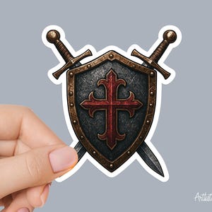 May include: A sticker featuring a shield with a red cross and two crossed swords. The shield is black with a bronze border and rivets. The swords have bronze hilts and silver blades. The sticker has a white border and is held by a hand.