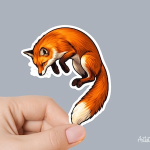 May include: A sticker featuring a stylized illustration of a jumping fox. The fox has orange fur with white and black accents. The sticker is held by a hand against a gray background. The artist's signature is in the bottom right corner.