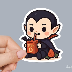 May include: A sticker featuring a cartoon vampire with black hair and a cape, drinking from a juice box labeled "TYPE O." The vampire is sitting next to a strawberry. The sticker has a white border.