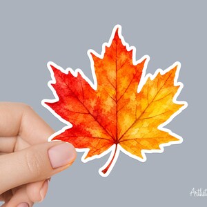 May include: A die-cut sticker shaped like a maple leaf featuring a watercolor-style gradient that transitions from deep red at the left side to vibrant orange and bright yellow on the right side, finished with a thin white border outline.