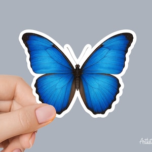 May include: A sticker featuring a vibrant blue butterfly with black and brown accents. The butterfly has a white border and is held by a hand against a gray background. The sticker is a design by ArtthetDesign.