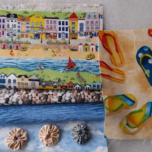 Może przedstawiać: Zestaw resztek tkaniny przedstawiający scenę plażową z kolorowymi domami, błękitnym oceanem i klapkami. Zestaw zawiera również dwie wstążki niebiesko-białe, wstążkę żółtą i trzy materiałowe rozety.