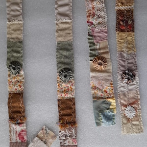 Può includere: Quattro strisce di tessuto con varie trame e colori, tra cui stampe floreali, pizzo e tessuti a tinta unita. Ogni striscia ha un elemento decorativo, come un fiore di stoffa o un bottone. Le strisce sono etichettate con testo scritto a mano.