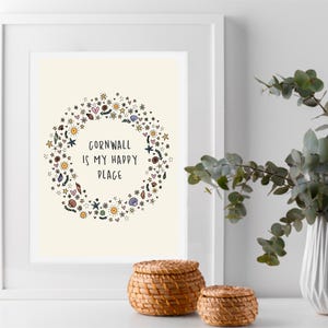 May include: Framed art print with the text "Cornwall is my happy place" surrounded by a floral wreath. Two woven baskets and a white vase with greenery are in the foreground.