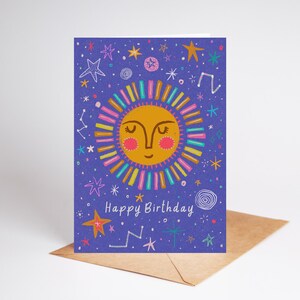 Puede incluir: Tarjeta de felicitación de cumpleaños con un sol sonriente sobre fondo morado, adornada con estrellas y la inscripción "Happy Birthday". El sol está rodeado de rayos de colores. La tarjeta está sobre un sobre marrón.
