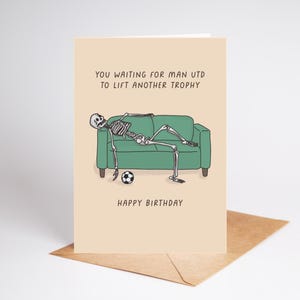 May include: A humorous birthday card featuring a skeleton lounging on a green couch with a soccer ball. The card reads "You waiting for Man Utd to lift another trophy" and "Happy Birthday". The card is on a brown envelope.