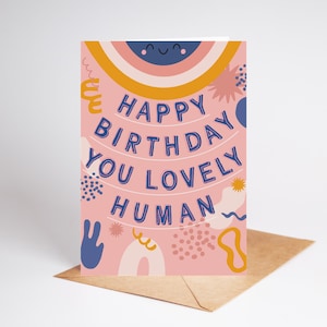 May include: A birthday card with the message "Happy Birthday You Lovely Human" in blue, arched text. The card features a pink background with decorative elements and a smiling rainbow. It sits on a brown envelope.
