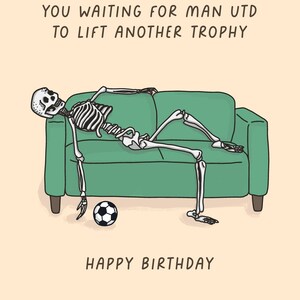 May include: A humorous birthday card featuring a cartoon skeleton lounging on a green couch with a soccer ball. The card includes the text "YOU WAITING FOR MAN UTD TO LIFT ANOTHER TROPHY" and "HAPPY BIRTHDAY" on a beige background.