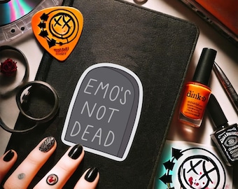 Könnte beinhalten: Ein schwarzes Notizbuch mit einem Aufkleber "EMO'S NOT DEAD". Ein orangefarbener Gitarrenplektrum mit schwarzem Design befindet sich neben dem Aufkleber. Nagellack, ein Ring und eine CD sind ebenfalls sichtbar.