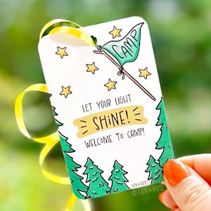 May include: A white rectangular tag with a green and yellow design. The tag features a flag that says "CAMP", stars, and text that reads "LET YOUR LIGHT SHINE! WELCOME TO CAMP!" A yellow ribbon is attached.