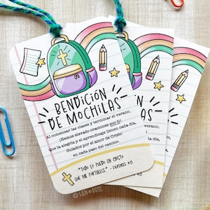 Op de afbeelding: Drie witte papieren labels met een rugzak-, potlood- en regenboogontwerp. De labels hebben de tekst "BENDICIÓN DE MOCHILAS" en een bijbelvers. De labels zijn vastgebonden met blauw en groen touw.