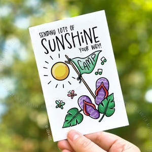 以下が含まれることがあります： 「Sending lots of sunshine your way!」のテキストが入った白いグリーティングカード。黄色い太陽、Campと書かれた緑の旗、紫色のビーチサンダル、蝶、音符が描かれています。カードは手で持たれています。
