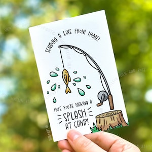 Könnte beinhalten: Eine weiße Grußkarte mit Angelmotiv. Die Karte zeigt eine Cartoon-Illustration einer Angelrute mit einem Fisch und Wassertröpfchen. Der Text auf der Karte lautet: "Sending a line from home!" und "Hope you're making a splash at camp!"