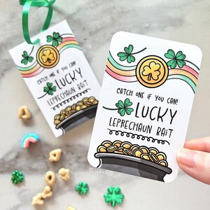 May include: Two white rectangular tags with green ribbon, featuring a pot of gold, shamrocks, and rainbow accents. Text reads "Catch one if you can! Lucky Leprechaun Bait." Scattered around are cereal pieces and shamrock candies.