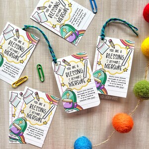 May include: White gift tags with the text "YOU ARE A BLESSING BEYOND MEASURE" and illustrations of a backpack, pencil, and ruler. The tags have a rainbow and star design, and are tied with blue and green twine.