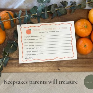 May include: A card with the text "Wishes for Our Little Cutie" and lines for writing, surrounded by oranges and eucalyptus. The card sits on a wooden board. The image also includes the text "Keepsakes parents will treasure".