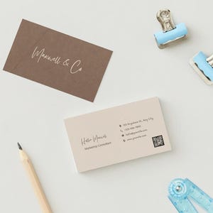 Op de afbeelding: Een witte visitekaartje met de tekst "Hollie Williams, Marketing Consultant" en contactgegevens, waaronder een adres, telefoonnummer, e-mailadres, websiteadres en een QR-code. De kaart ligt op een wit oppervlak met een bruin visitekaartje met de tekst "Maxwell & Co" in witte schrijfletters.