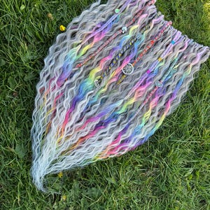 May include: A set of long, wavy, gray synthetic hair extensions with rainbow colored streaks and silver charms. The extensions are laid out on a bed of green grass.