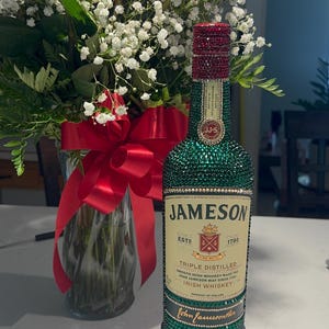 May include: A green Jameson Irish Whiskey bottle, embellished with sparkling green and red crystals. The bottle features the Jameson label and the text "1780". A bouquet of red roses and white flowers is in the background.