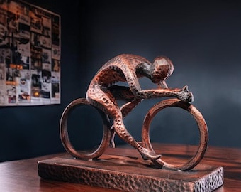 Puede incluir: Una escultura de bronce de un ciclista en posición de carrera, montada sobre una base rectangular. La escultura tiene una superficie texturizada y está colocada sobre un fondo oscuro. El ciclista está inclinado hacia adelante, con las manos en el manillar.