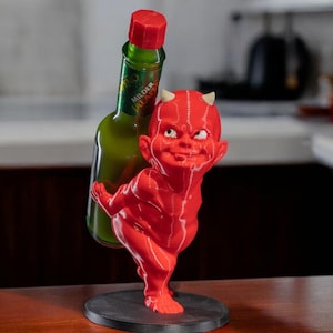 May include: A red devil figurine holding a green glass bottle with a red cap. The bottle has a green label with white text. The devil has small white horns and is standing on a black circular base. The background is blurred.