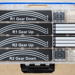 May include: A clear plastic tackle box with black labels that say "R1 Gear Down", "R1 Gear Up", "R2 Gear Up", and "R2 Gear Down". The labels have white letters and lines. The box has two small clear compartments with black labels that say "A, B, C" and "D, E, F".