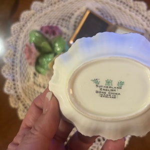 May include: Close-up of the bottom of a white porcelain dish with a scalloped edge. The dish has a green and white floral design and the text "Sutherland English Bone China Made in England" printed on the bottom.