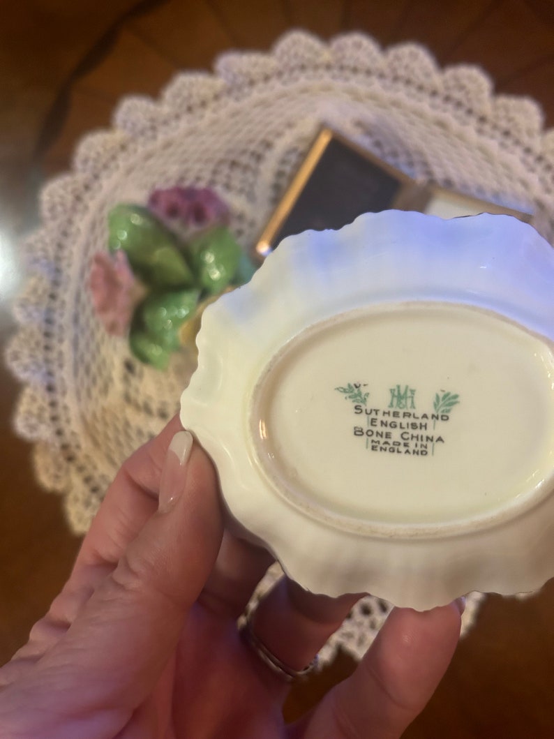 May include: Close-up of the bottom of a white porcelain dish with a scalloped edge. The dish has a green and white floral design and the text "Sutherland English Bone China Made in England" printed on the bottom.