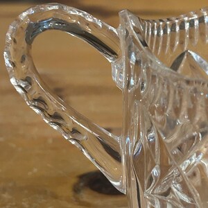 Op de afbeelding: Een heldere glazen roomkan met een geslepen glasontwerp en een handvat. De roomkan is gemaakt van kristal en heeft een delicaat, ingewikkeld patroon.
