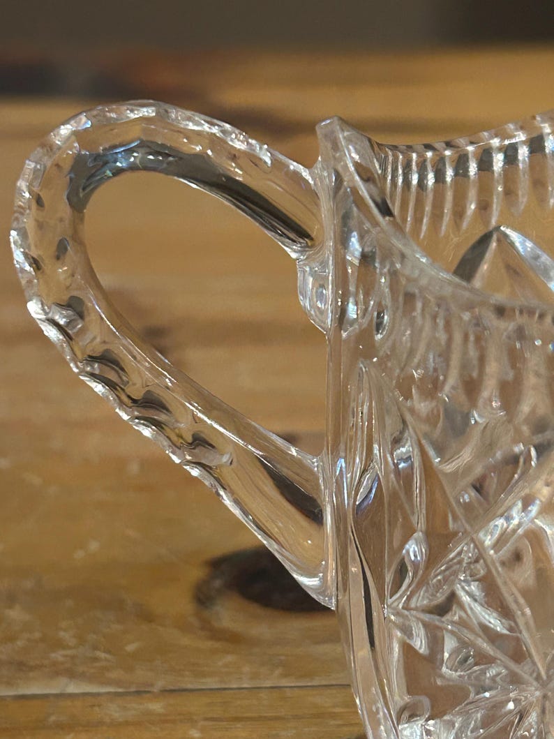 Op de afbeelding: Een heldere glazen roomkan met een geslepen glasontwerp en een handvat. De roomkan is gemaakt van kristal en heeft een delicaat, ingewikkeld patroon.