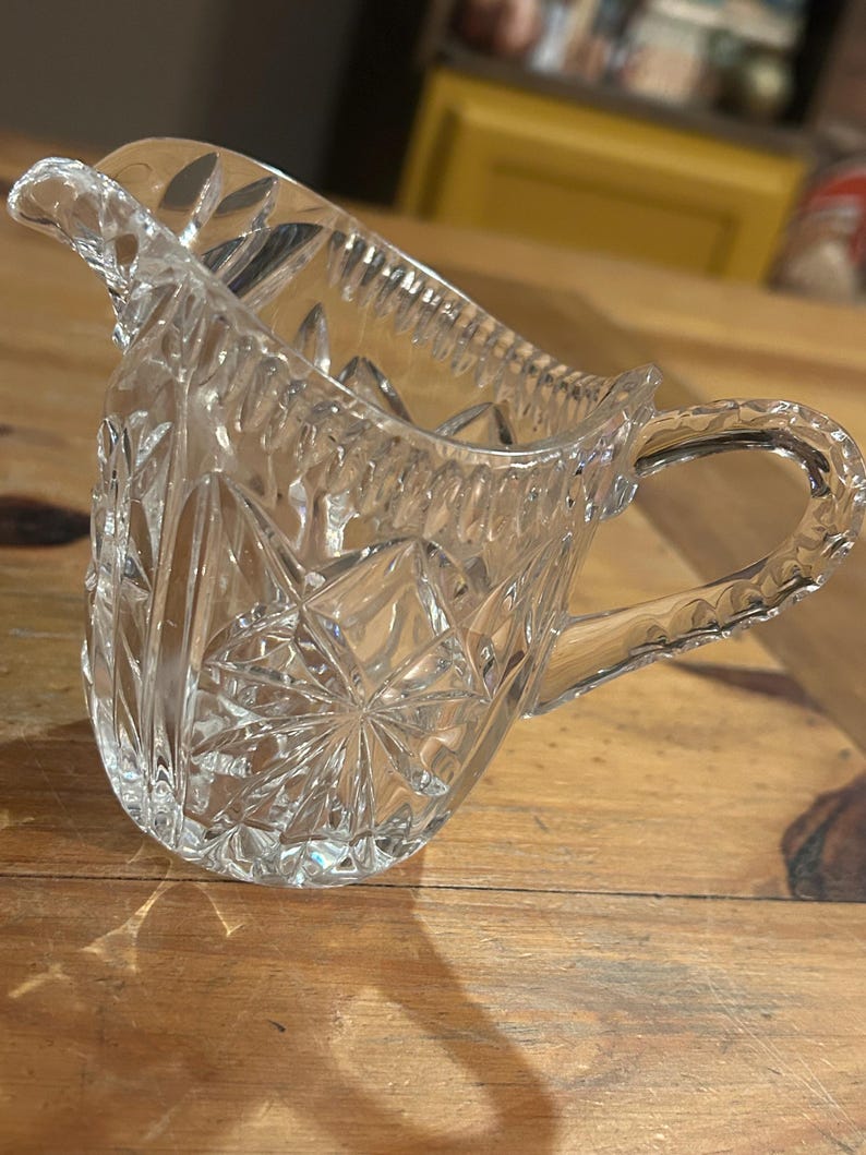 Op de afbeelding: Een heldere glazen roomkan met een geslepen glasontwerp. De roomkan heeft een handvat en een tuit.