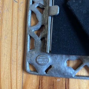 May include: A silver picture frame with an ornate design. The frame has a black velvet backing and a metal latch. The frame is stamped with the text "1883 Kirk & Sille Pewter".