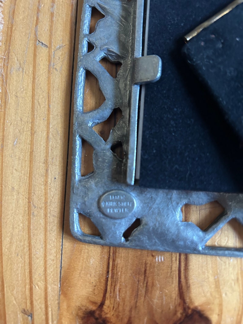 May include: A silver picture frame with an ornate design. The frame has a black velvet backing and a metal latch. The frame is stamped with the text "1883 Kirk & Sille Pewter".