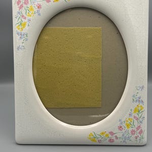 May include: White ceramic picture frame with an oval opening and a square outer shape. The frame is decorated with pastel-colored floral accents in pink, yellow, and blue. The frame has a crackle finish.