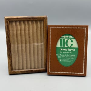 May include: Two vintage photo frames. One is a rectangular wooden frame with a brown finish. The other is a brown leather-like frame with the text "photo frame by Intercraft" and the dimensions 3 1/4 x 4 1/4 overall - 2 x 3 opening.
