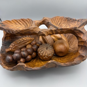 May include: A carved wooden fruit bowl with a leaf-like design. The bowl contains carved wooden grapes, a pumpkin, a pear, and other fruits. The wood has a natural brown color with darker accents in the carved details. The bowl is a decorative piece.